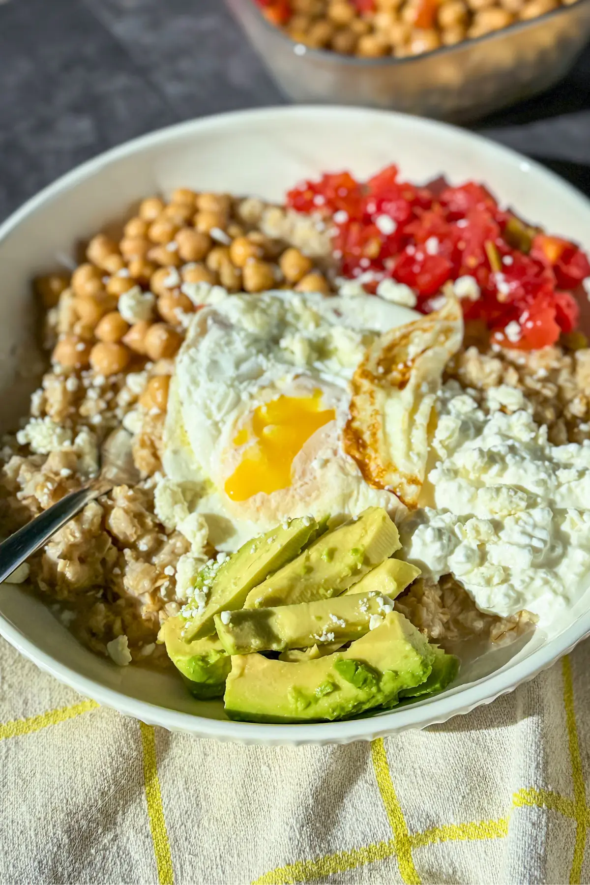 A fully assembled high protein savory Mediterranean-inspired bowl of oatmeal with avocado slices, tomatoes, chickpeas, cottage cheese, and a runny egg.