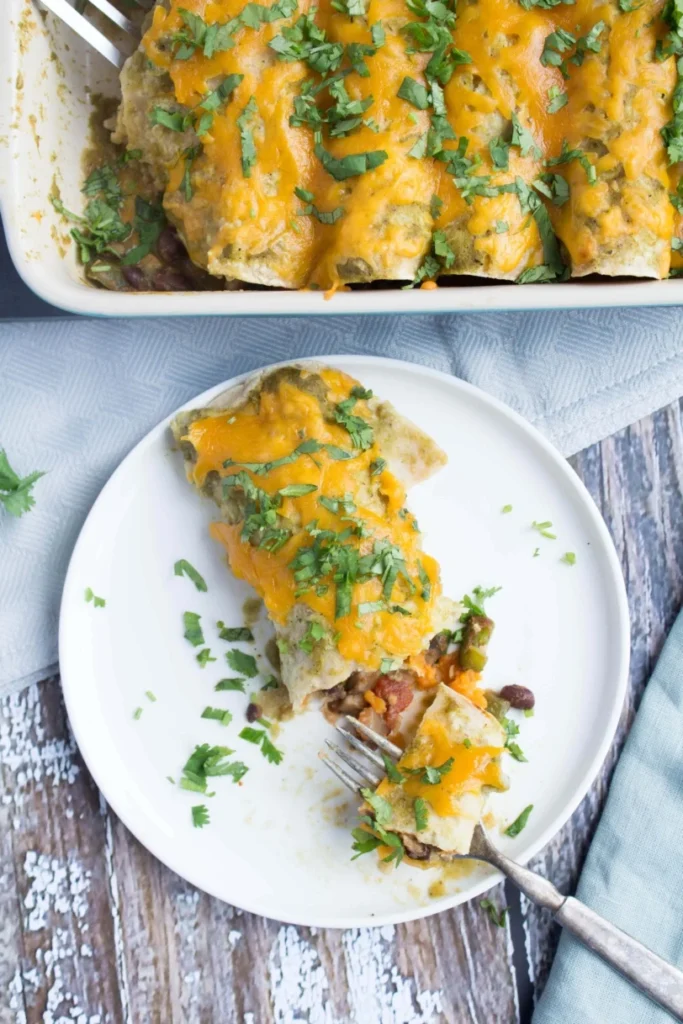 A sweet potato and black bean enchilada on a plate with a fork on a table next to a pan of enchiladas.