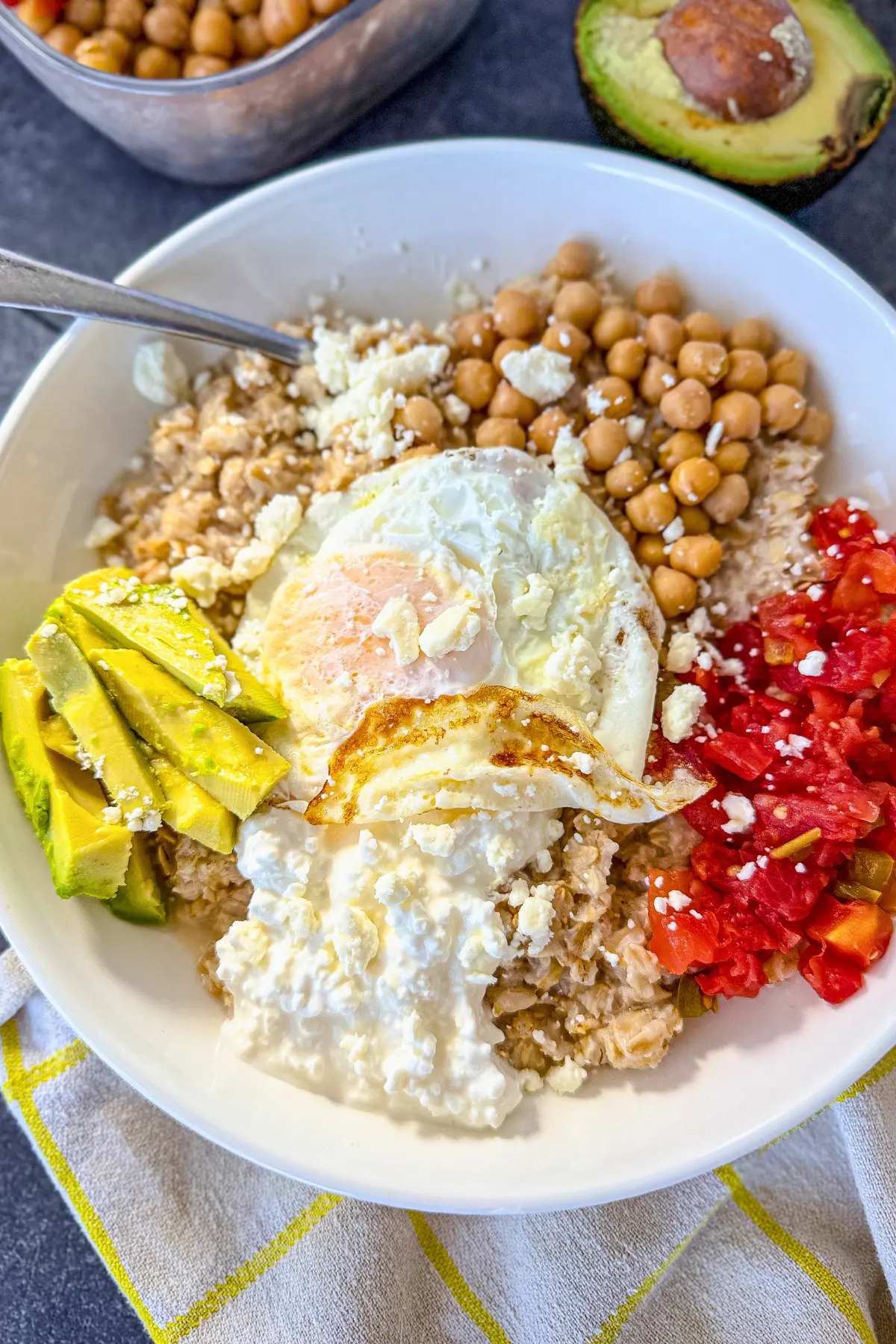 A savory high protein oatmeal bowl with cottage cheese, chickpeas and tomatoes on a counter next to a halved avocado.