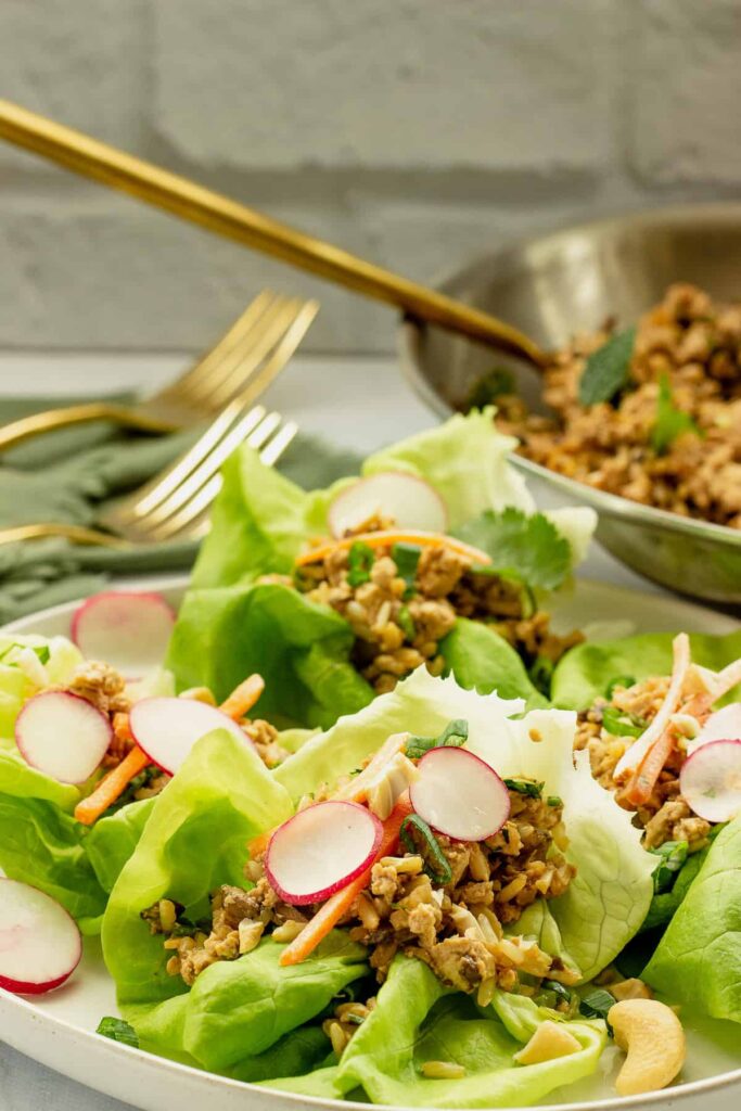 Four tofu lettuce wraps on a plate on a table with a pan of tofu in the background.