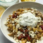 A bowl of pasta with feta cheese, sun-dried tomatoes, and topped with a garlic yogurt on a counter next to lemon wedges and a bowl of yogurt.
