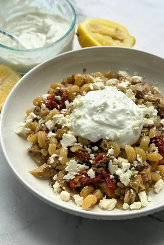 A bowl of pasta with feta cheese, sun-dried tomatoes, and topped with a garlic yogurt on a counter next to lemon wedges and a bowl of yogurt.