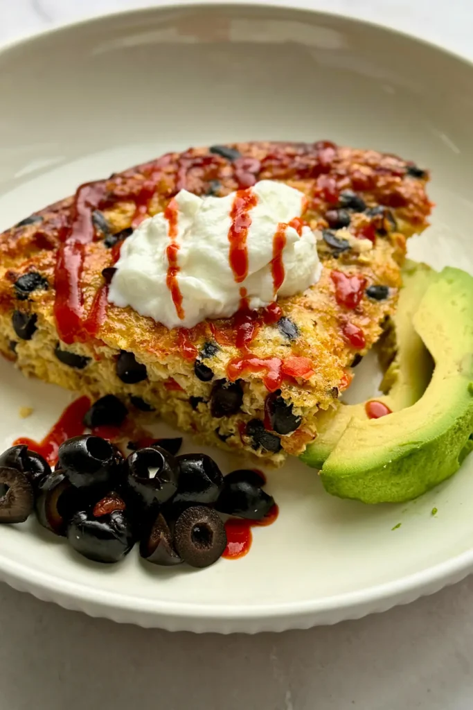 A piece of the quinoa breakfast bake on a plate next to olives, avocado and topepd with hot sauce and yogurt.
