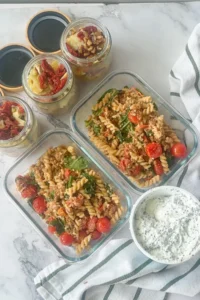 Two meal prep containers with lentil feta pasta, a bowl of cottage cheese with chia seeds and three salad jars with chickpeas and artichokes in them.