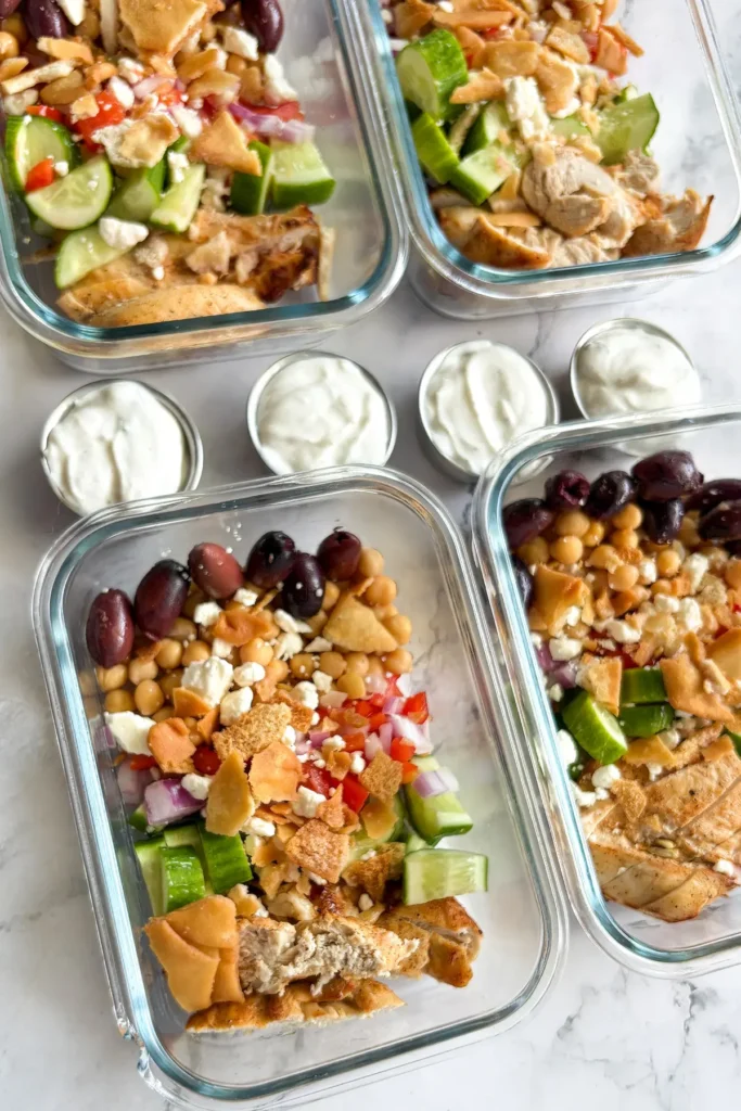 Meal prepped Greek chicken bowls on a counter next to small containers of the garlic Greek yogurt.