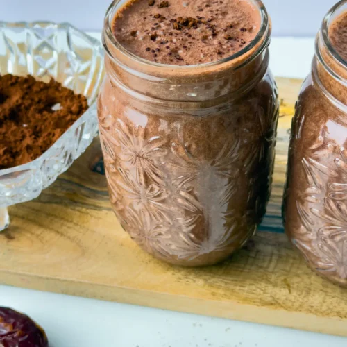 Two glasses full of the dark chocolate collagen smoothie on a wooden cutting board on a counter next to a bowl of cacao powder and dates.