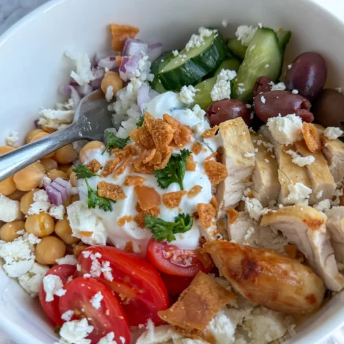 Two fully assembled Greek chicken protein bowls on a table topped with garlic yogurt and crushed pita chips.