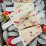 High protein birthday cake popsicles stacked on ice cubes on a cookie sheet next to fresh strawberries.