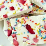 Slices of high protein cottage cheese bark with sprinkles, strawberries and white chocolate chips stacked on a plate.