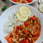 The crockpot coconut chicken with tomato sauce, carrots, chickpeas, and rice on two plates garnished with lemon wedges on a table.