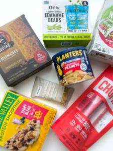A bunch of healthy, portable, store-bought snacks with protein on a counter - like protein bars, edamame beans, and beef sticks.