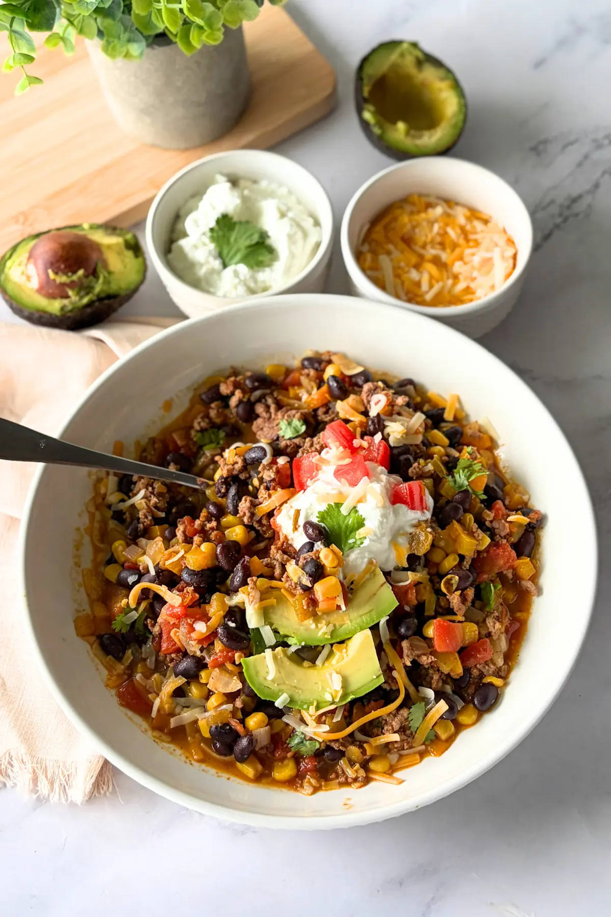 A bowl of one pot chili on a counter next to bowls of cheese adn Greek yogurt and an avocado.