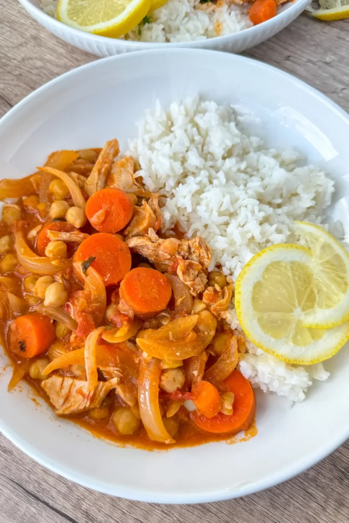 A close-up of the creamy coconut milk and tomato sauce chicken with chickpeas, carrots, rice, and garnished with lemons on a plate.