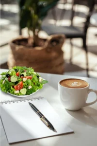 A cup of coffee next to a pad of paper and a pen and a small salad on a table.