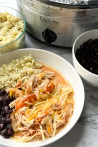 A plate of salsa chicken with rice and black beans next to a bowl of black beans, a bowl of rice, and a crockpot with salsa chicken in it on a counter.