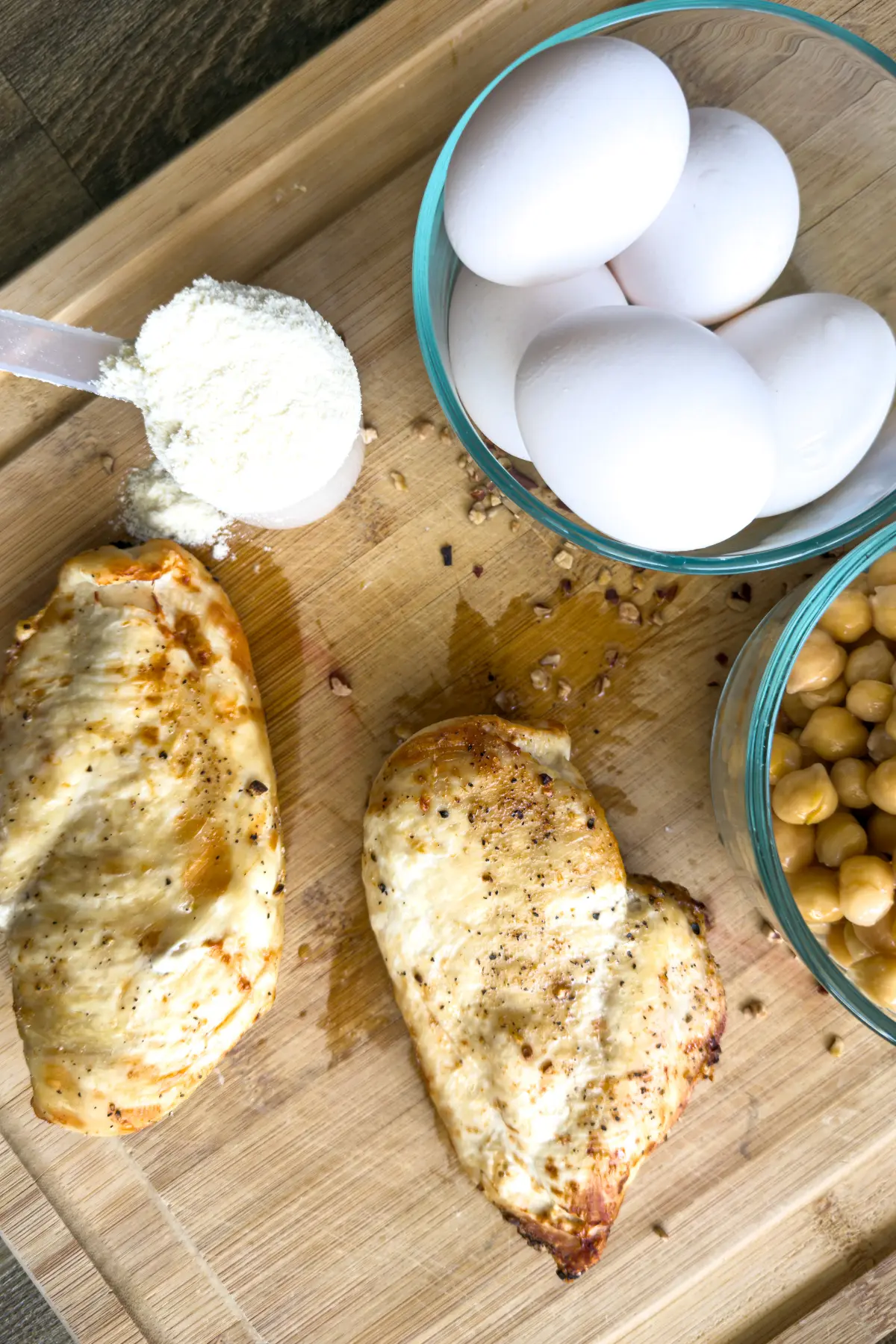 Chopped grilled chicken on a cutting board next to a bowl of chickpeas, a scoop of protein powder, and a bowl of eggs.