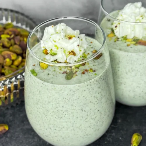 Two cottage cheese pistachio pudding smoothies in glasses topped with whipped cream and crushed pistachios on a counter next to a bowl of pistachios.