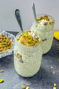 Two large mason jars filled with pistachio pudding overnight oats and topped with whipped cream, pistachios, and banana slices on a counter next to a bowl of pistachios.