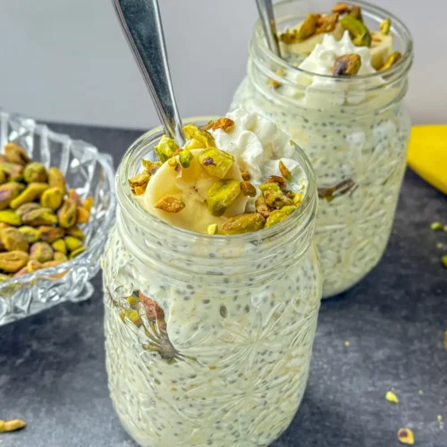 Two large mason jars filled with pistachio pudding overnight oats and topped with whipped cream, pistachios, and banana slices on a counter next to a bowl of pistachios.