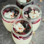 Four mini cups of high protein vanilla pudding topped with whipped cream, nuts, and mixed berries on a counter.