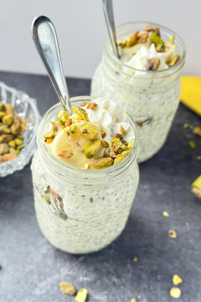 Two large mason jars with pistachio pudding overnight oats with spoons, topped with whipped cream, pistachios, and banana slices.