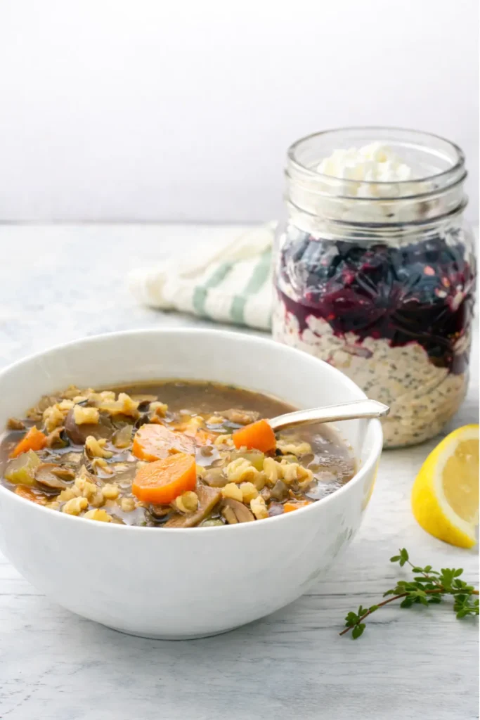 A bowl of barley soup next to lemon kefir overnight oats with berries on a table next to a lemon wedge.