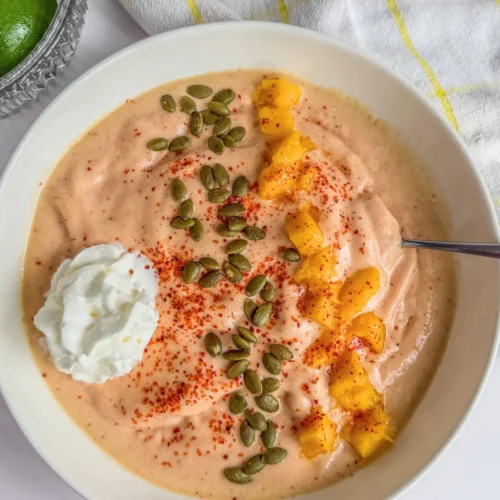 A tropical smoothie bowl topped with diced mango, whipped cream, pepitas and additional Tajin seasoning with a spoon in it on a table next to a yellow and white striped towel.