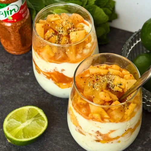 Two fully assembled tropical parfaits in glasses topped with diced pineapple, mango, and Tajin on a counter next to a bunch of mint and Tajin seasoning bottle.