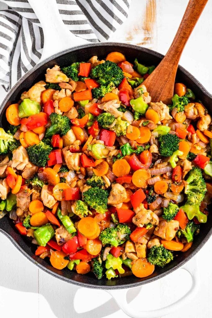 A birds-eye view of a pan of stir-fried veggies with chicken with a wooden spoon on a towel on a table.