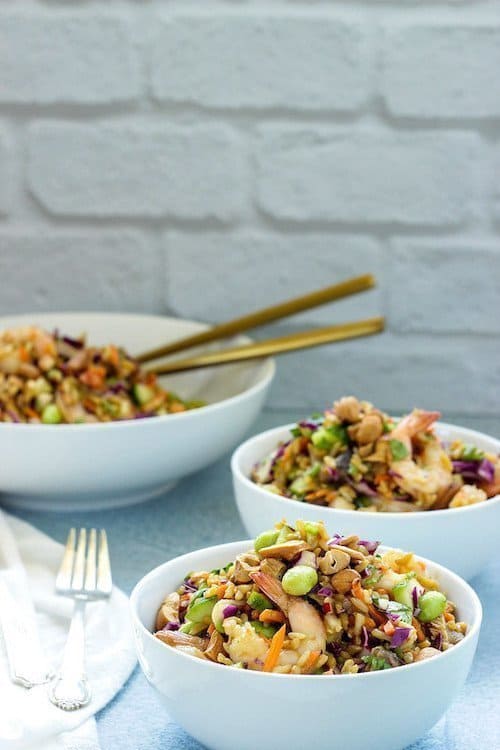 Two small bowls of Asian rice salad with shrimp next to a larger bowl with serving spoons in it on a counter.