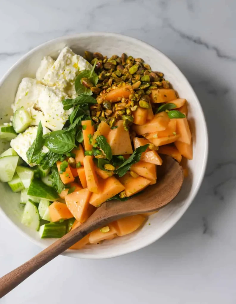 A birds-eye view of a bowl of cantaloupe salad with feta cheese and pistachios with a wooden spoon on a counter.