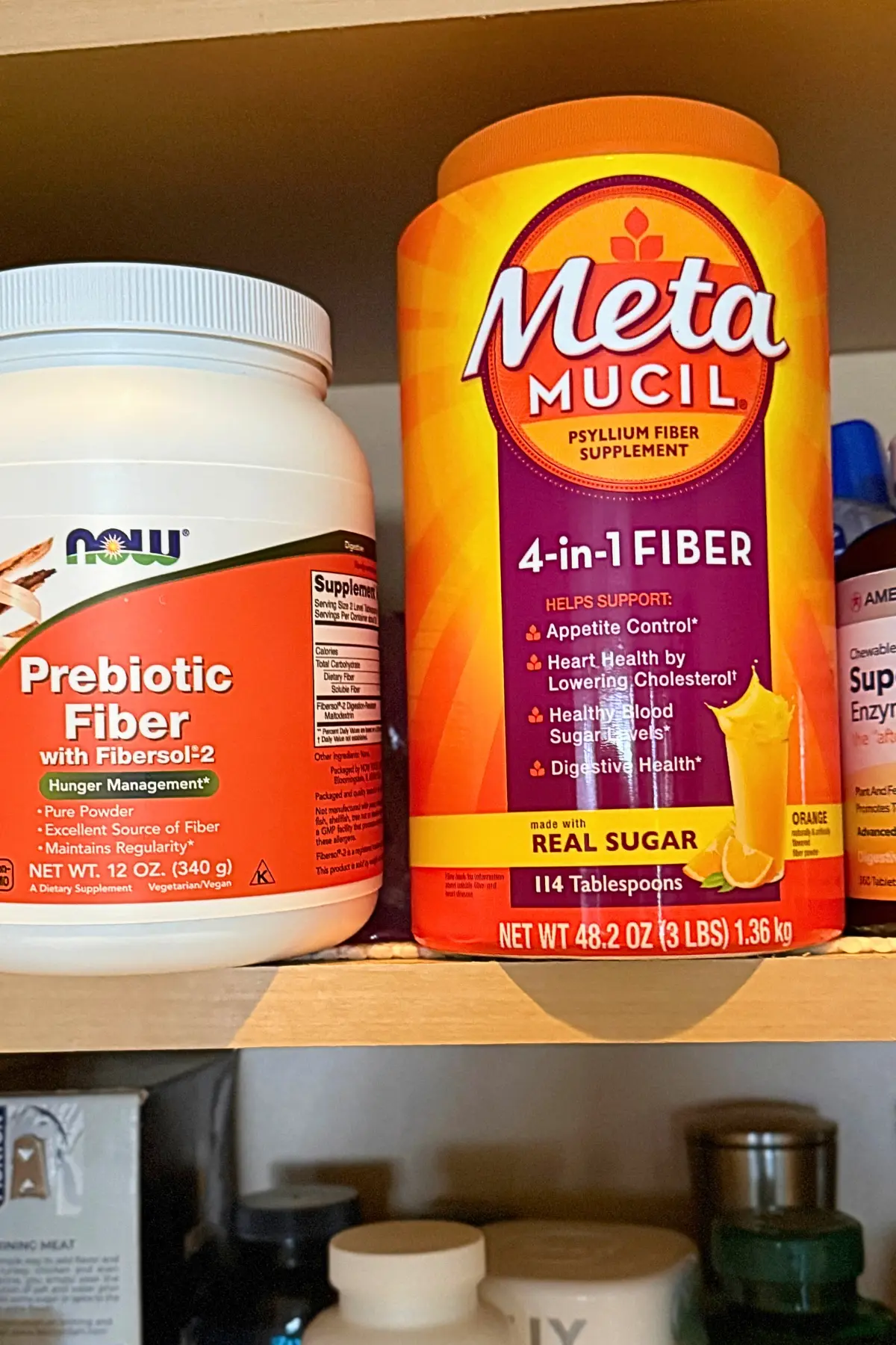 Two fiber supplements (Metamucil and Fibersol) on a shelf in a cupboard next to other supplements.