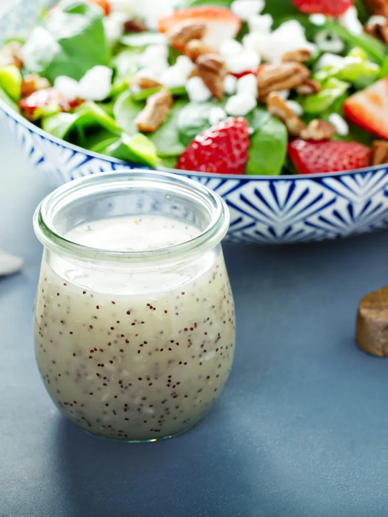 A strawberry grilled chicken salad in a bowl next to a glass of poppyseed dressing on a table.