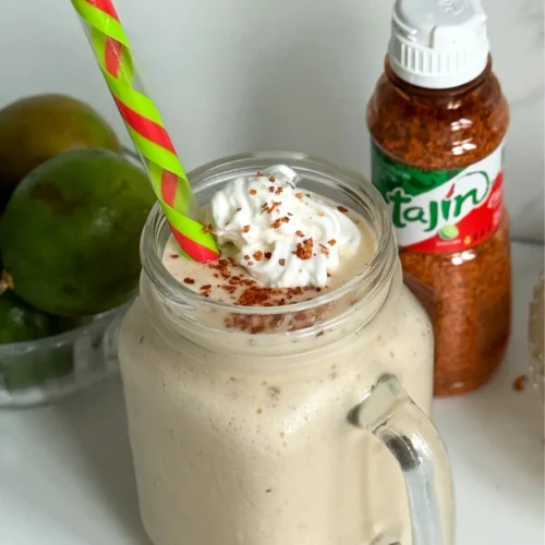 A tropical yogurt smoothie with Tajin seasoning in a glass topped with whipped cream and a colorful straw on a counter next to a bottle of Tajin seasoning.