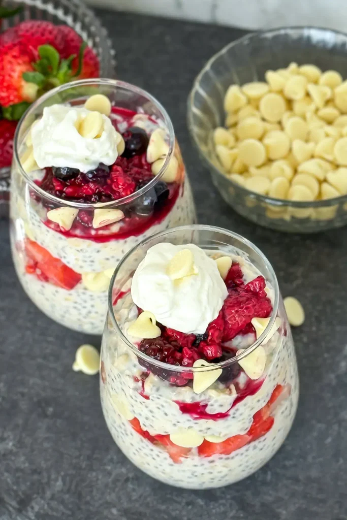Two white chocolate pudding overnight oats in glasses topped with raspberries, white chocolate chips and whipped cream on a counter.