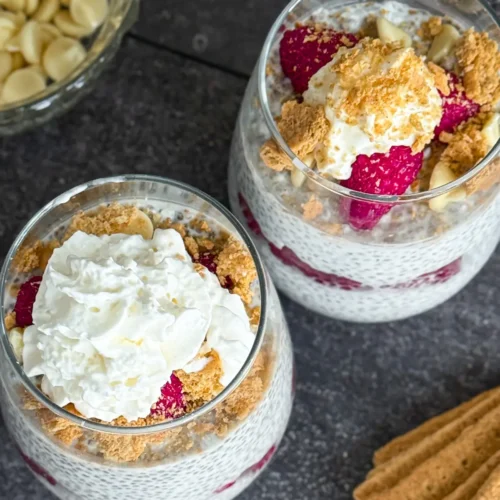 A birds-eye vie of two raspberry cheesecake chia puddings in glass topped with whipped cream and white chocolate chips on a counter next to a bowl of white chocolate.