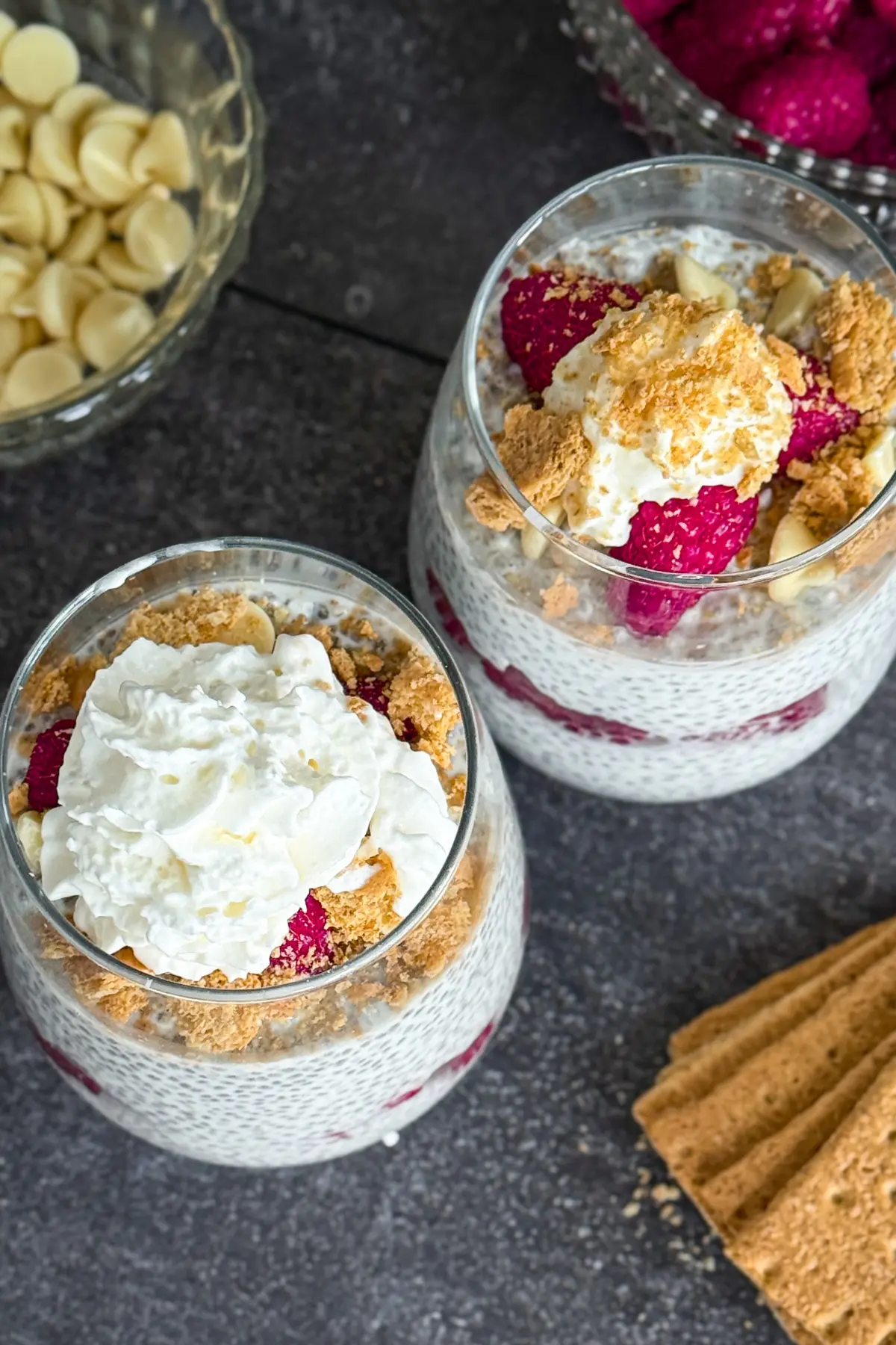 A birds-eye vie of two raspberry cheesecake chia puddings in glass topped with whipped cream and white chocolate chips on a counter next to a bowl of white chocolate.