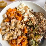 A close-up of a fully assembled butternut squash and chicken bowl drizzled with a maple and tahini dressing on a counter next to lemon halves and two bowls.
