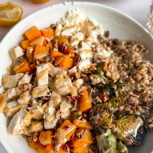 A close-up of a fully assembled butternut squash and chicken bowl drizzled with a maple and tahini dressing on a counter next to lemon halves and two bowls.