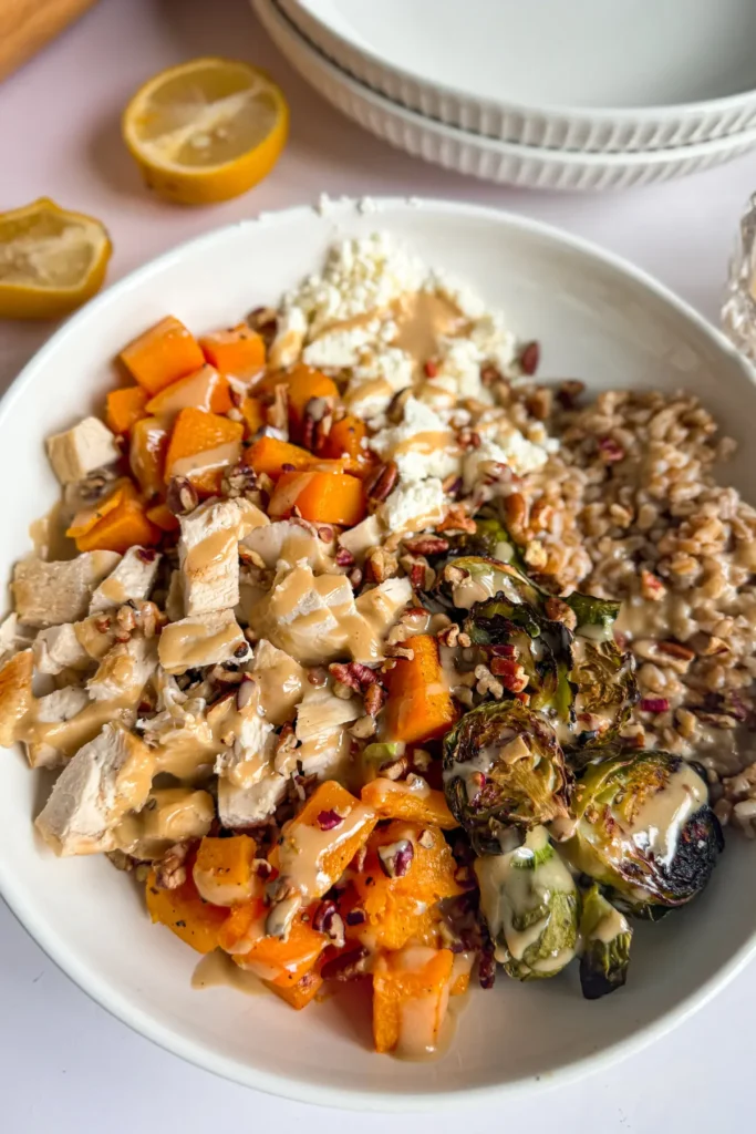 A close-up of a fully assembled butternut squash and chicken bowl drizzled with a maple and tahini dressing on a counter next to lemon halves and two bowls.