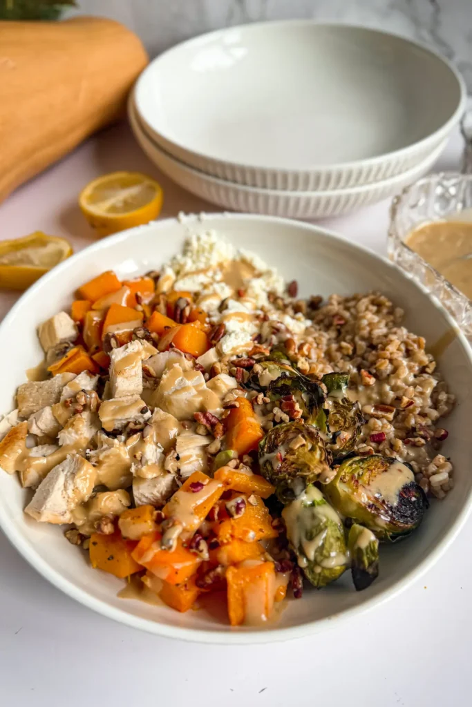 A fully assembled butternut squash harvest bowl drizzled in maple tahini on a counter next to a butternut squash and two bowls.
