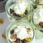 Small mini pistachio pudding cups topped with whipped cream, dark chocolate chips and chopped pistachios on a counter.