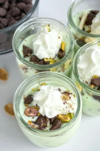 Small mini pistachio pudding cups topped with whipped cream, dark chocolate chips and chopped pistachios on a counter.