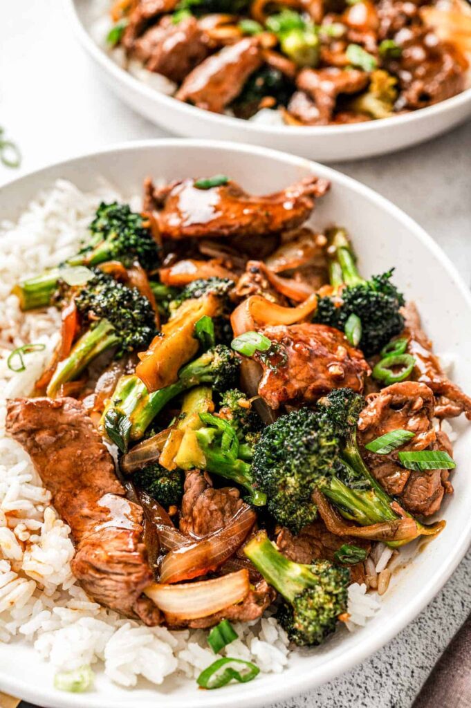 Two fully assembled beef and broccoli plates with rice on a table.