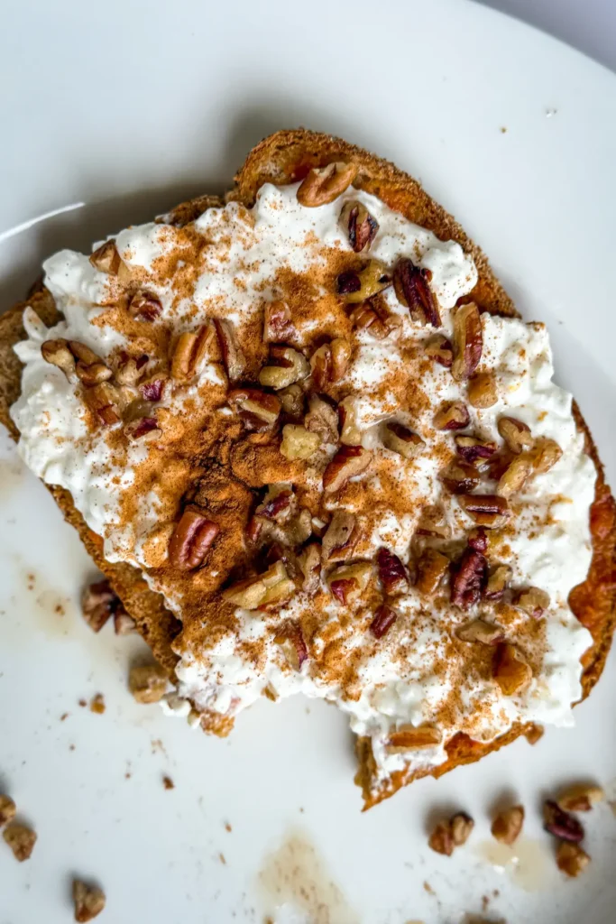 A slice of toast with pumpkin puree spread on it, cottage cheese, chopped pecans and maple syrup on a white plate.