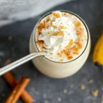 A birds-eye view of a creamy banana and cinnamon protein shake topped with whipped cream in a glass with a straw in it next to cinnamon sticks.