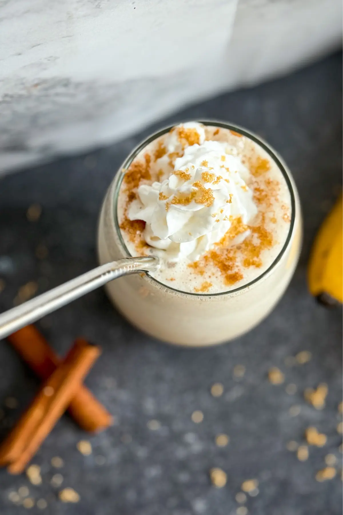 A birds-eye view of a creamy banana and cinnamon protein shake topped with whipped cream in a glass with a straw in it next to cinnamon sticks.