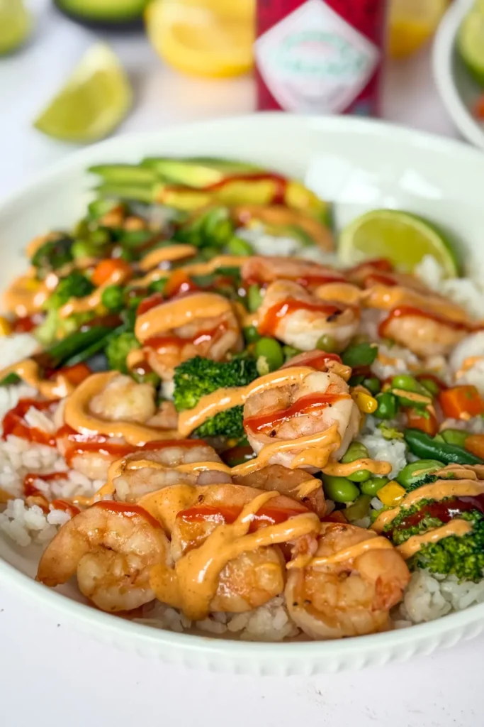 A close-up of a shrimp, rice and veggie bowl with sriracha and spicy mayo drizzled over it on a counter next to half a lime and different sauces.