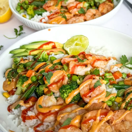Two fully assembled macro-friendly shrimp and rice bowls on a counter next to half an avocado, half a lime, and different sauces.
