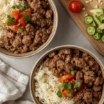 A birds-eye view of two rice and ground beef bowls on a counter next to a hand towel, a cutting board with chopped veggies on it.